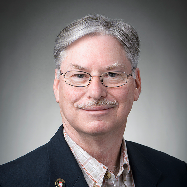 James Lattis, a white man, sits in a traditional sport coat plus label pin and checkered button up shirt. His hair is greying and he has a slight smile and narrow thin rimmed glasses.